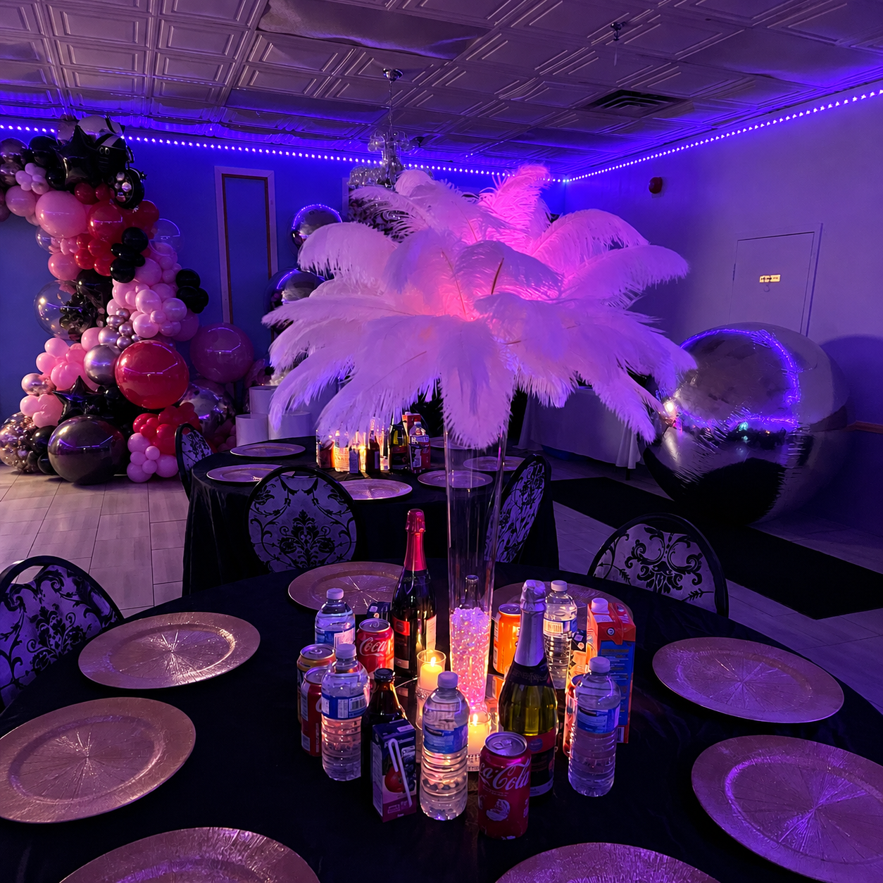 Sabreena’s Sweet Sixteen party table setup featuring a tall white feather centerpiece, purple ambient lighting, black table linens, silver place settings, and pink balloon decor.