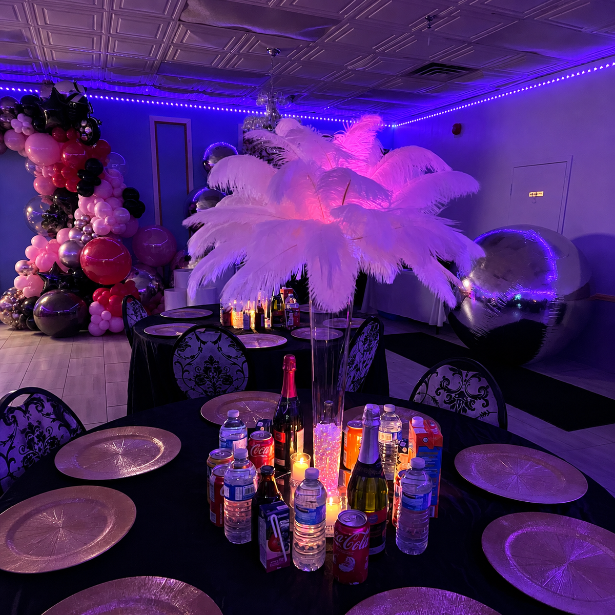 Sabreena’s Sweet Sixteen party table setup featuring a tall white feather centerpiece, purple ambient lighting, black table linens, silver place settings, and pink balloon decor.