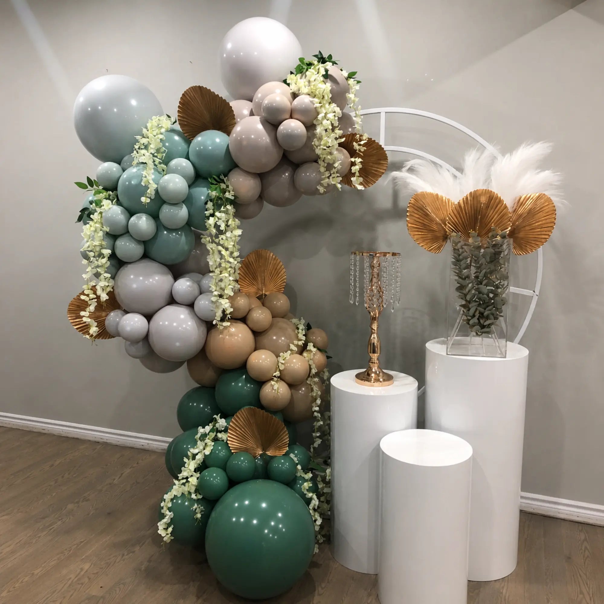 “Oh Baby” baby shower backdrop featuring sage green, white, and metallic gold balloon garlands, greenery and floral accents, a white circular backdrop, and decorative BABY blocks.