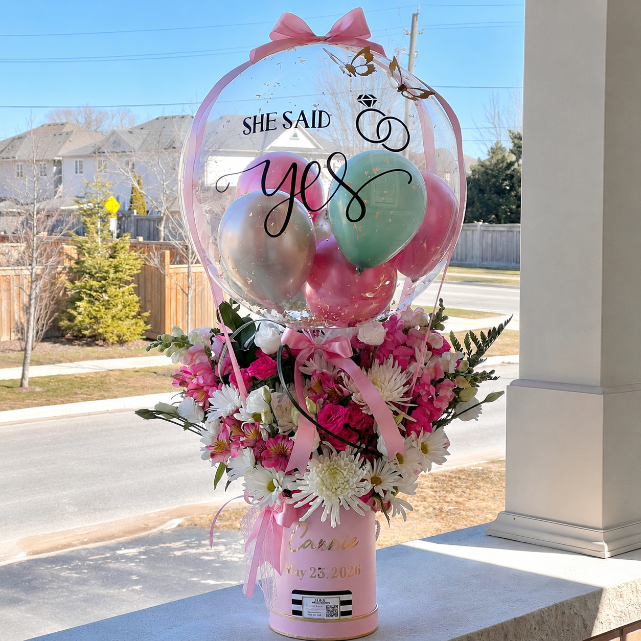 Engagement celebration arrangement featuring a personalised “She Said Yes” clear balloon, pink and green balloons, and pink and white flowers.