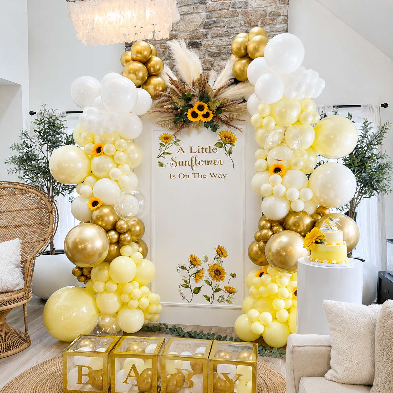 Sunflower baby shower backdrop featuring yellow, white, and gold balloon garlands, sunflower accents, a “A Little Sunflower Is On The Way” sign, baby blocks, cake pedestal, and a boho rattan chair.