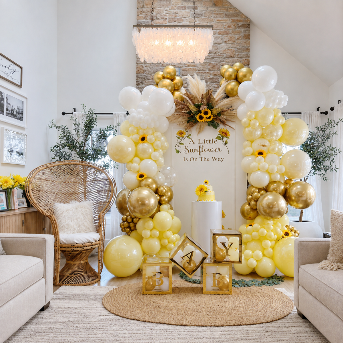 Sunflower baby shower backdrop featuring yellow, white, and gold balloon garlands, sunflower accents, a “A Little Sunflower Is On The Way” sign, baby blocks, cake pedestal, and a boho rattan chair.