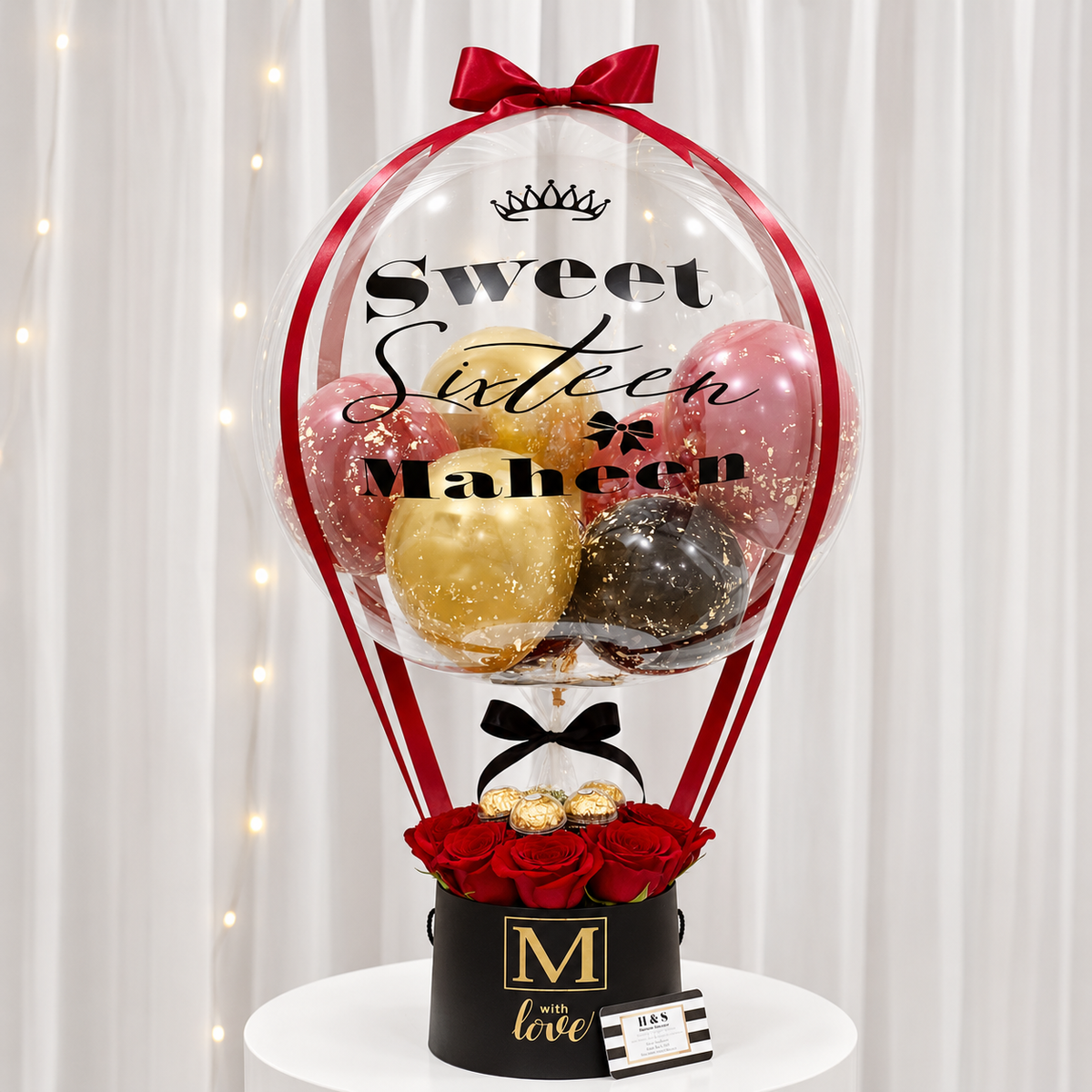 Sweet sixteen gift arrangement featuring a personalized clear balloon with red, gold, pink, and black balloons, red roses, chocolates, red ribbon accents, and a black gift box with gold detailing