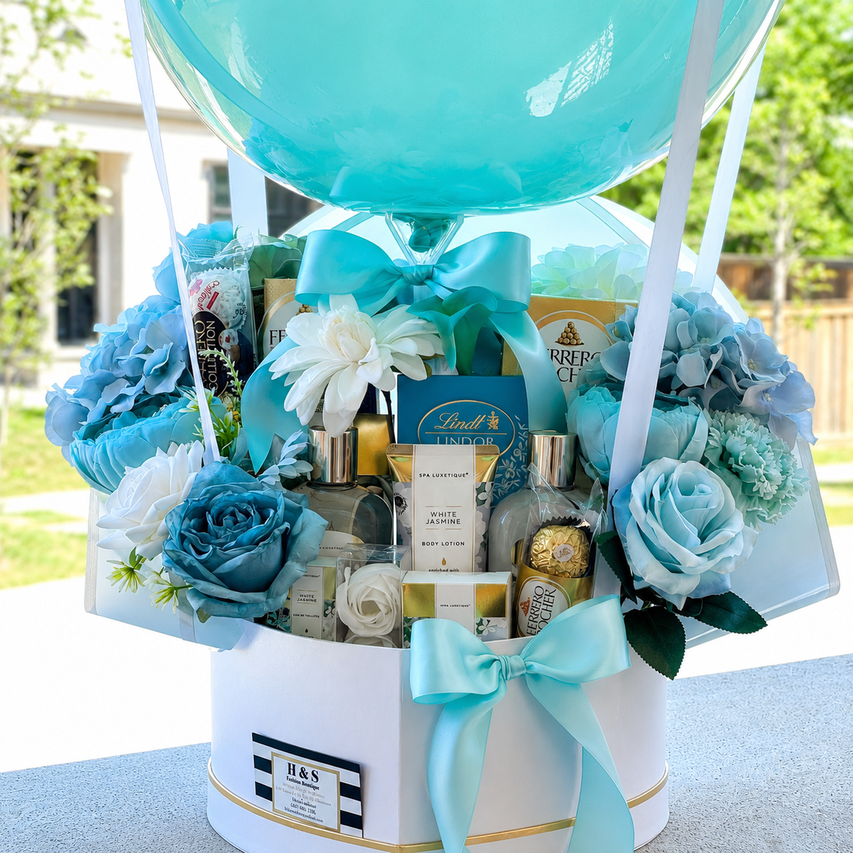 Close-up of a teacher appreciation gift arrangement featuring aqua and white flowers, Lindt and Ferrero Rocher chocolates, beauty products, and turquoise ribbon accents.