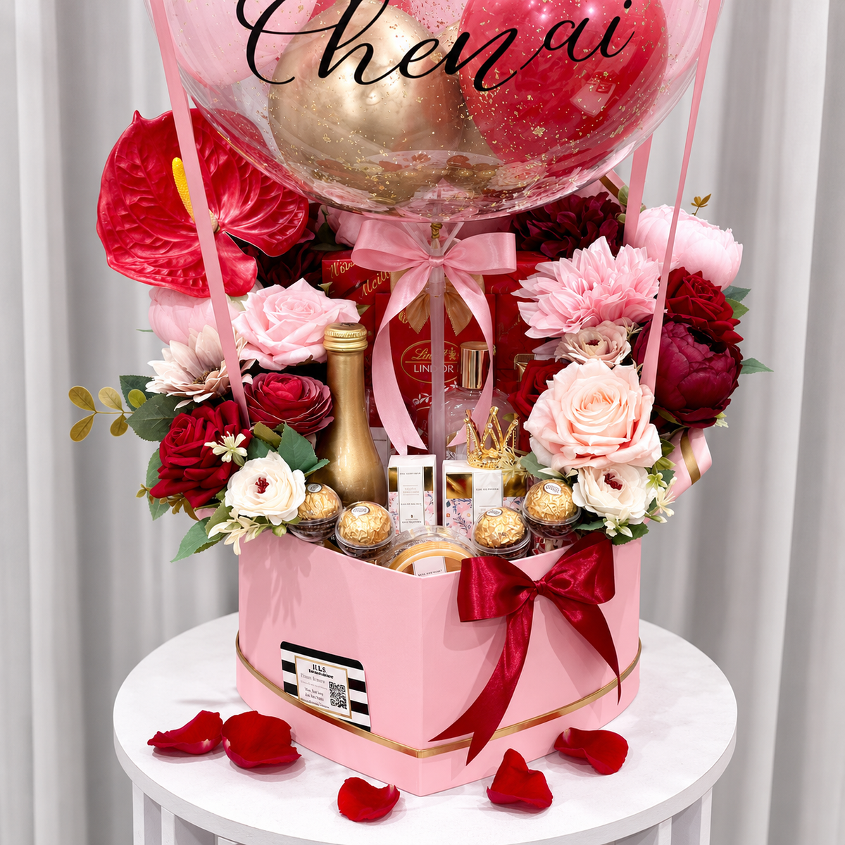 Close-up of a Valentine’s gift arrangement featuring red and pink flowers, chocolates, beauty products, a gold bottle, pink ribbons, and a clear balloon.