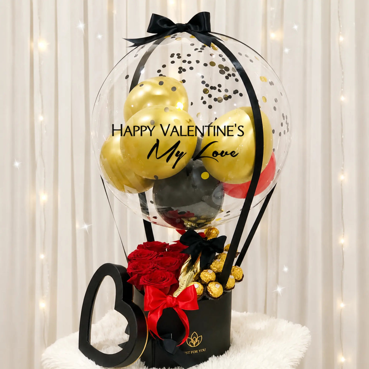 Valentine’s Day gift arrangement featuring a personalized clear balloon with gold, black, and red balloons, red roses, gold-wrapped chocolates, black and red ribbon accents, and a black gift box.