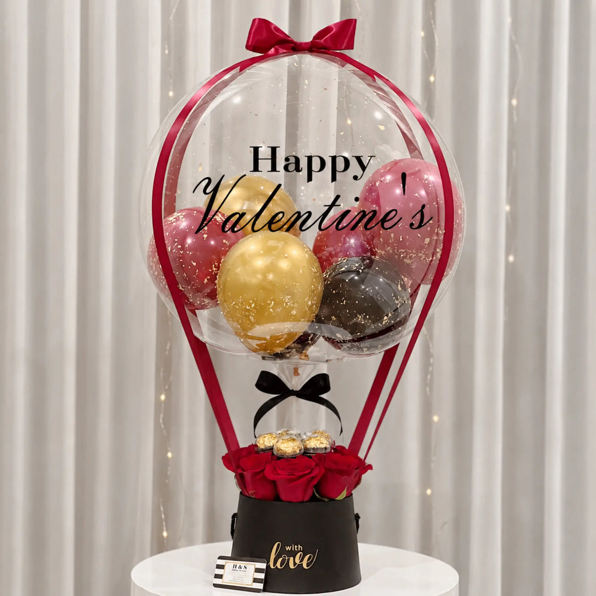Valentine’s Day gift arrangement featuring a personalized clear balloon with red, gold, and black balloons, red roses, gold-wrapped chocolates, black gift box, and red and black ribbon accents.
