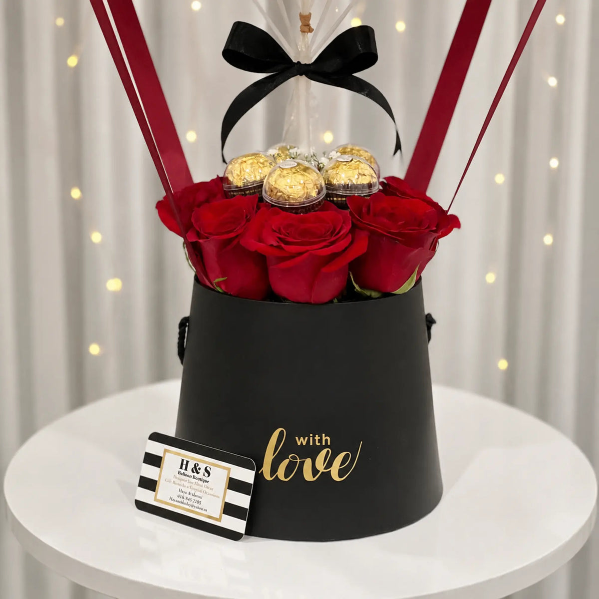 Close-up of a Valentine’s Day gift arrangement featuring red roses, gold-wrapped chocolates, a black “with love” gift box, black ribbon bow, and red balloon straps.