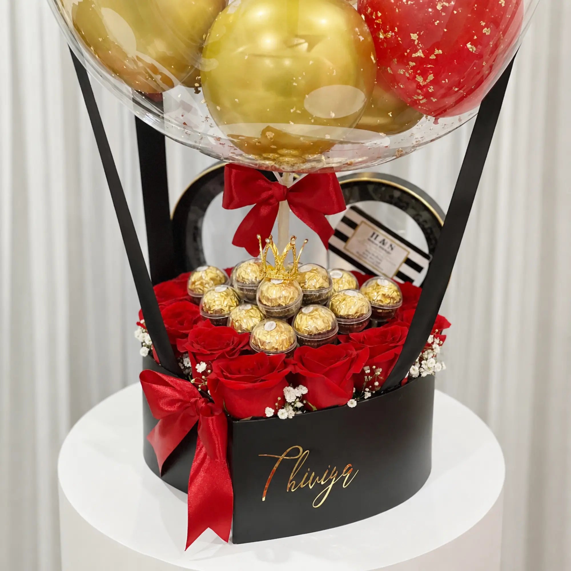 Valentine’s Day proposal gift arrangement featuring a personalized clear “Will You Be My Valentine?” balloon with gold, red, and black balloons, red roses, gold-wrapped chocolates, black gift box, and red ribbon accents.