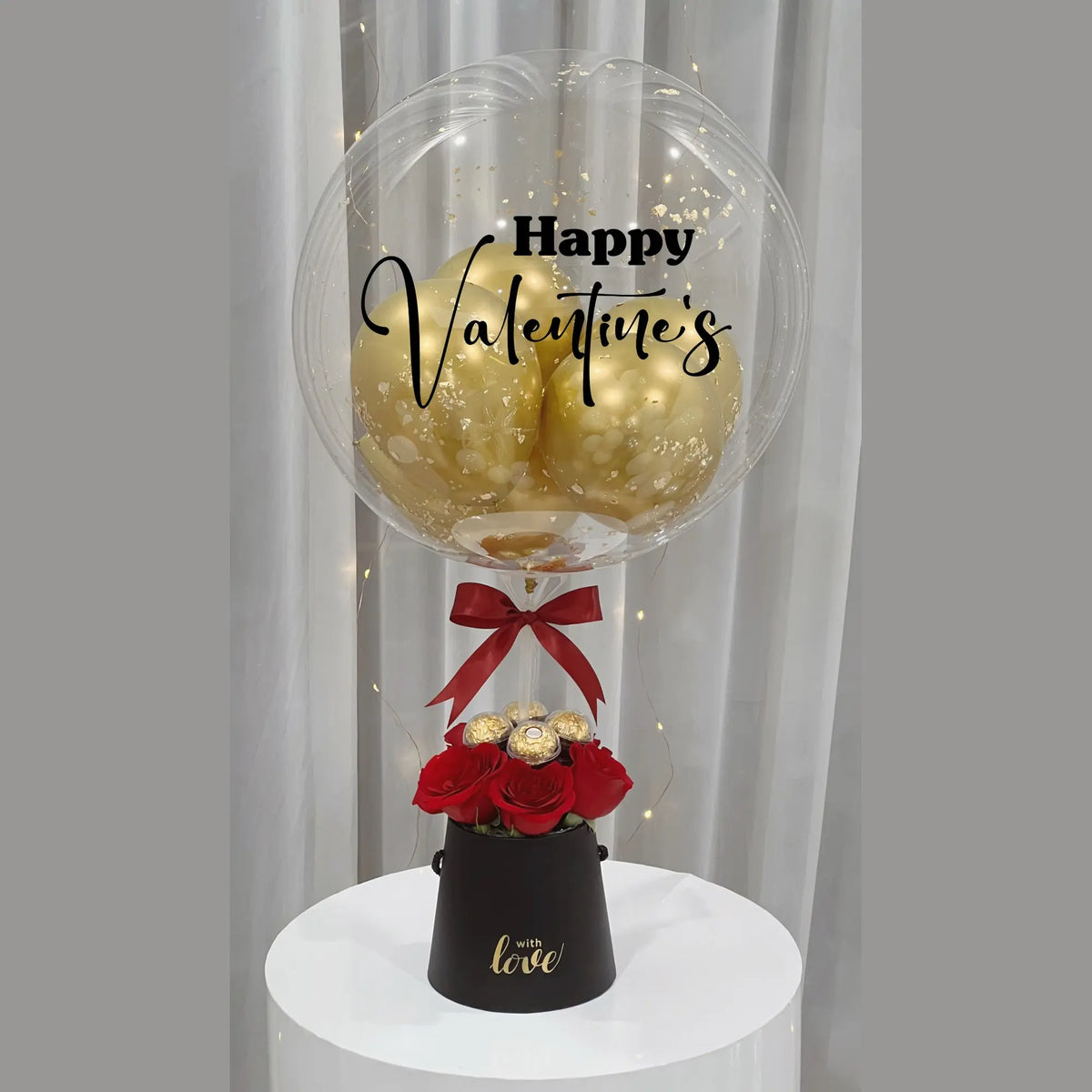 Valentine’s Day gift arrangement featuring a personalized clear balloon with gold balloons, red roses, gold-wrapped chocolates, a black gift box, and red ribbon accents.