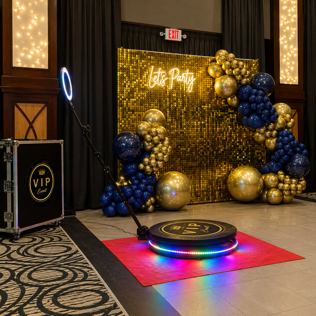 VIP event setup featuring a gold shimmer wall, navy blue and metallic gold balloon garlands, a glowing “Let’s Party” sign, and a 360 photo booth platform with ring light.