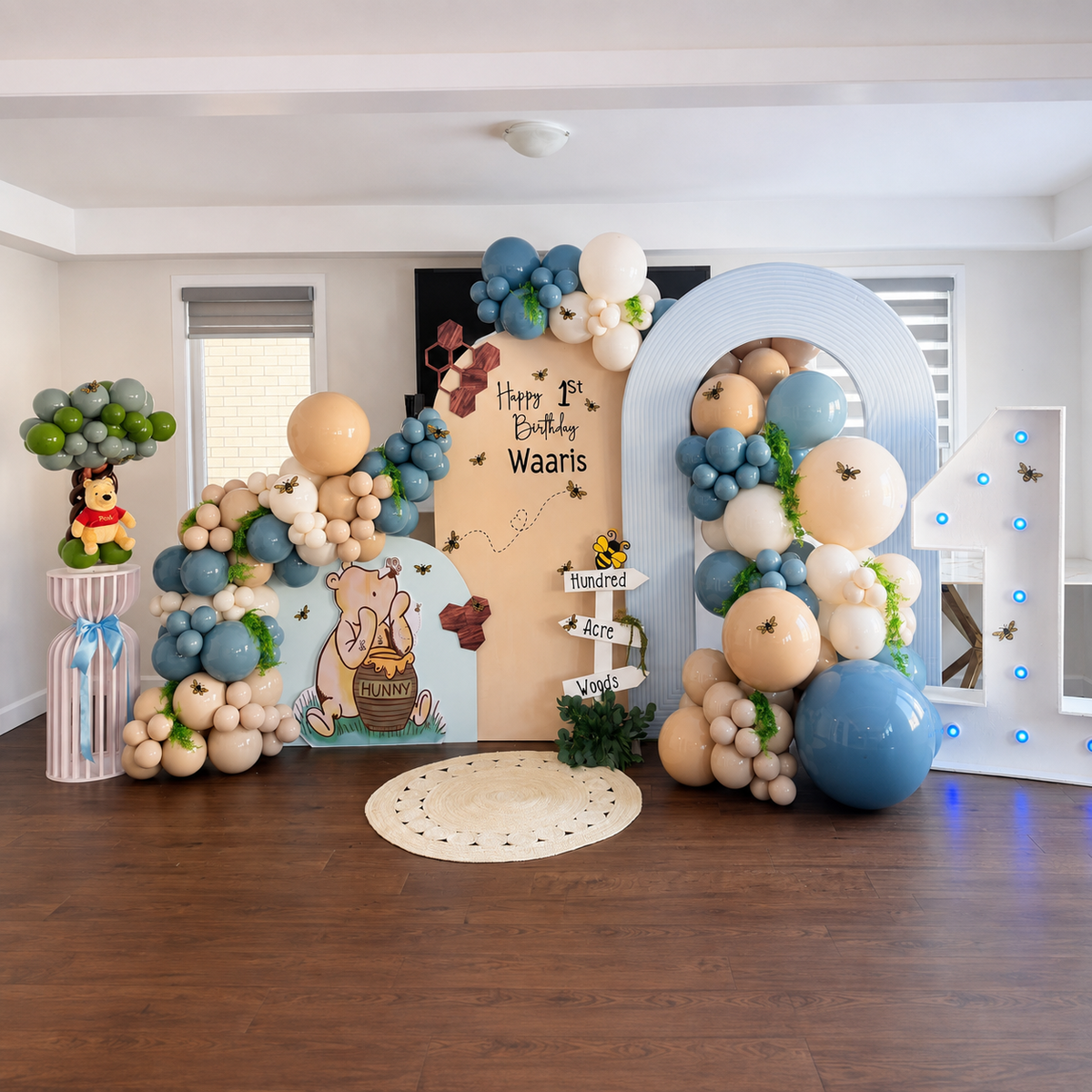 Waaris’ first birthday backdrop featuring a Winnie the Pooh theme, blue, beige, and white balloon garlands, honeybee decorations, and a large illuminated number one.