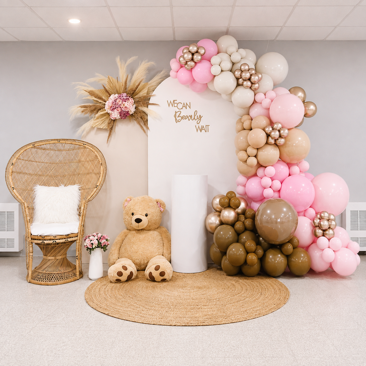 “We Can Bearly Wait” baby shower backdrop featuring pink, cream, tan, and brown balloon garlands, pampas grass and floral accents, a teddy bear, rattan peacock chair, white pedestal, and woven rug.