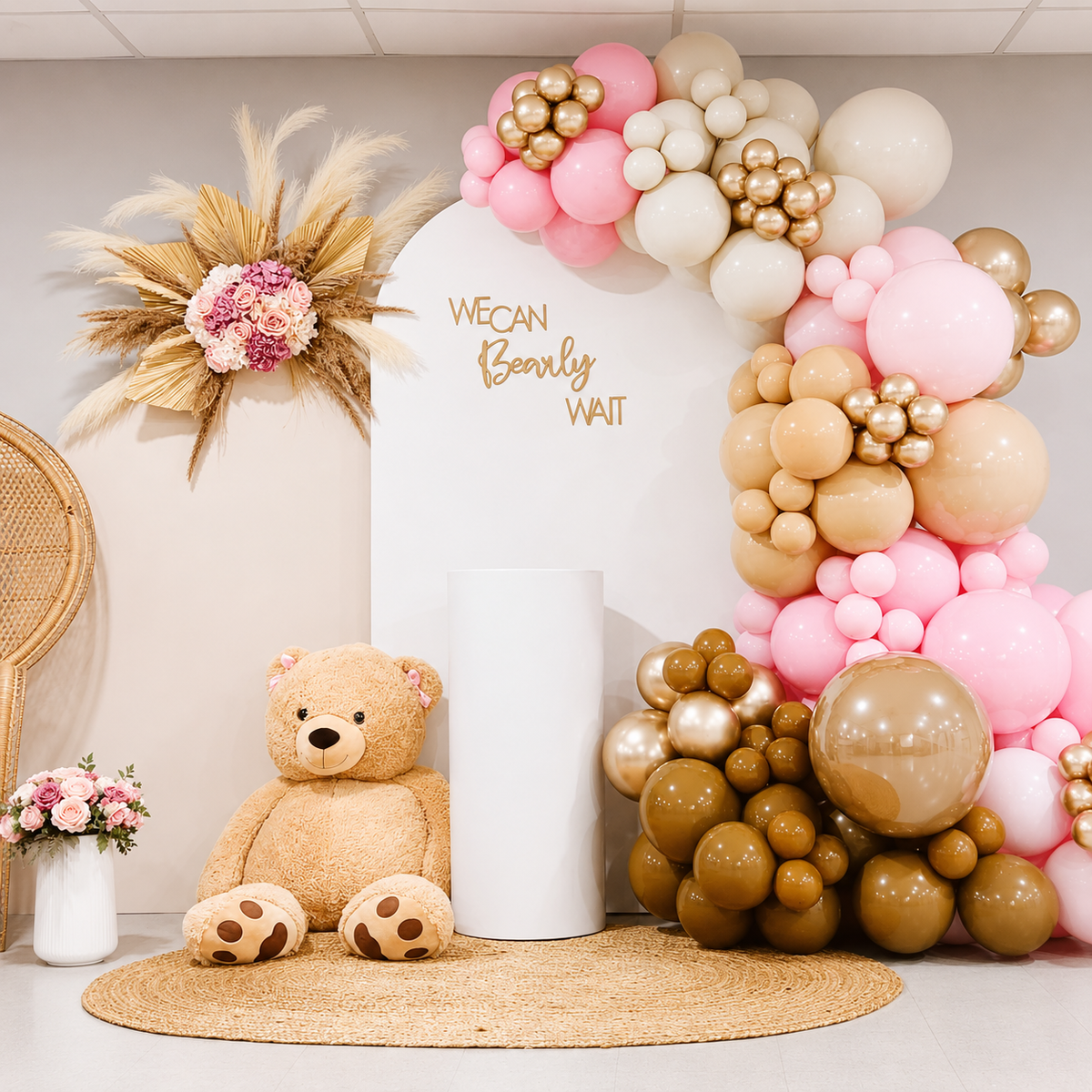 “We Can Bearly Wait” baby shower backdrop featuring pink, cream, tan, brown, and gold balloon garlands, a large teddy bear, pampas grass and floral accents, and a white pedestal on a woven rug.