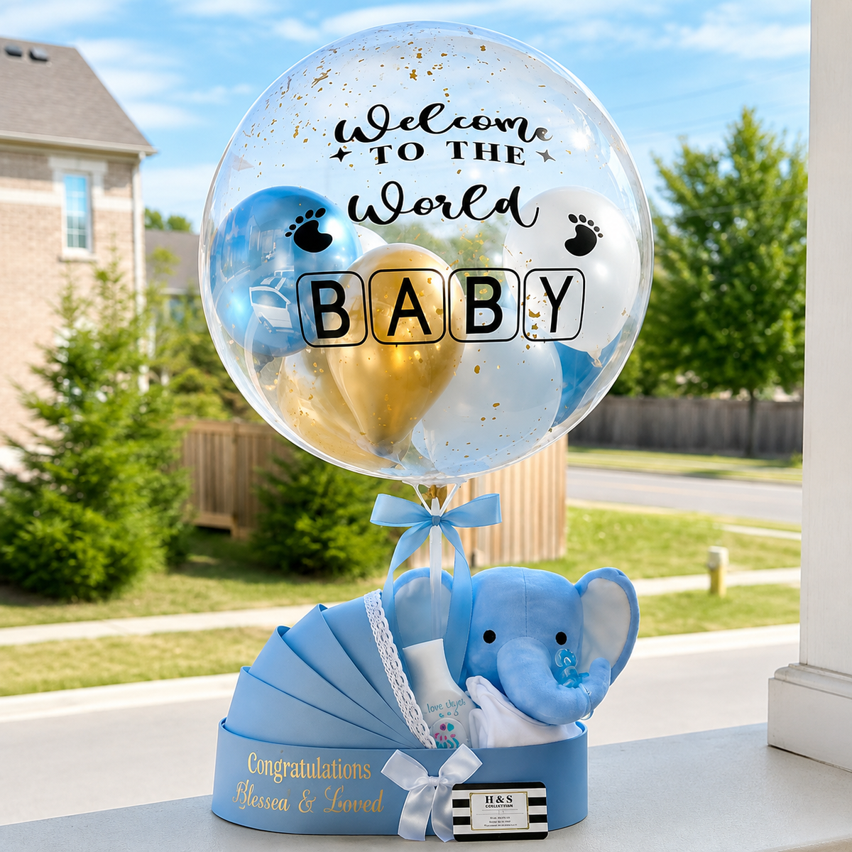 Welcome baby gift arrangement featuring a personalised clear balloon, blue, white, and gold balloons, a plush elephant, baby essentials, and blue ribbon accents.