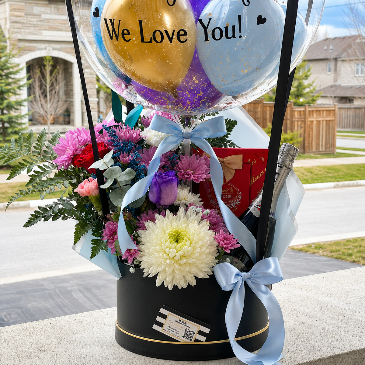 Close-up of a welcome to Canada gift arrangement featuring a personalised clear balloon, colorful flowers, chocolates, a sparkling beverage bottle, and light blue ribbon accents.