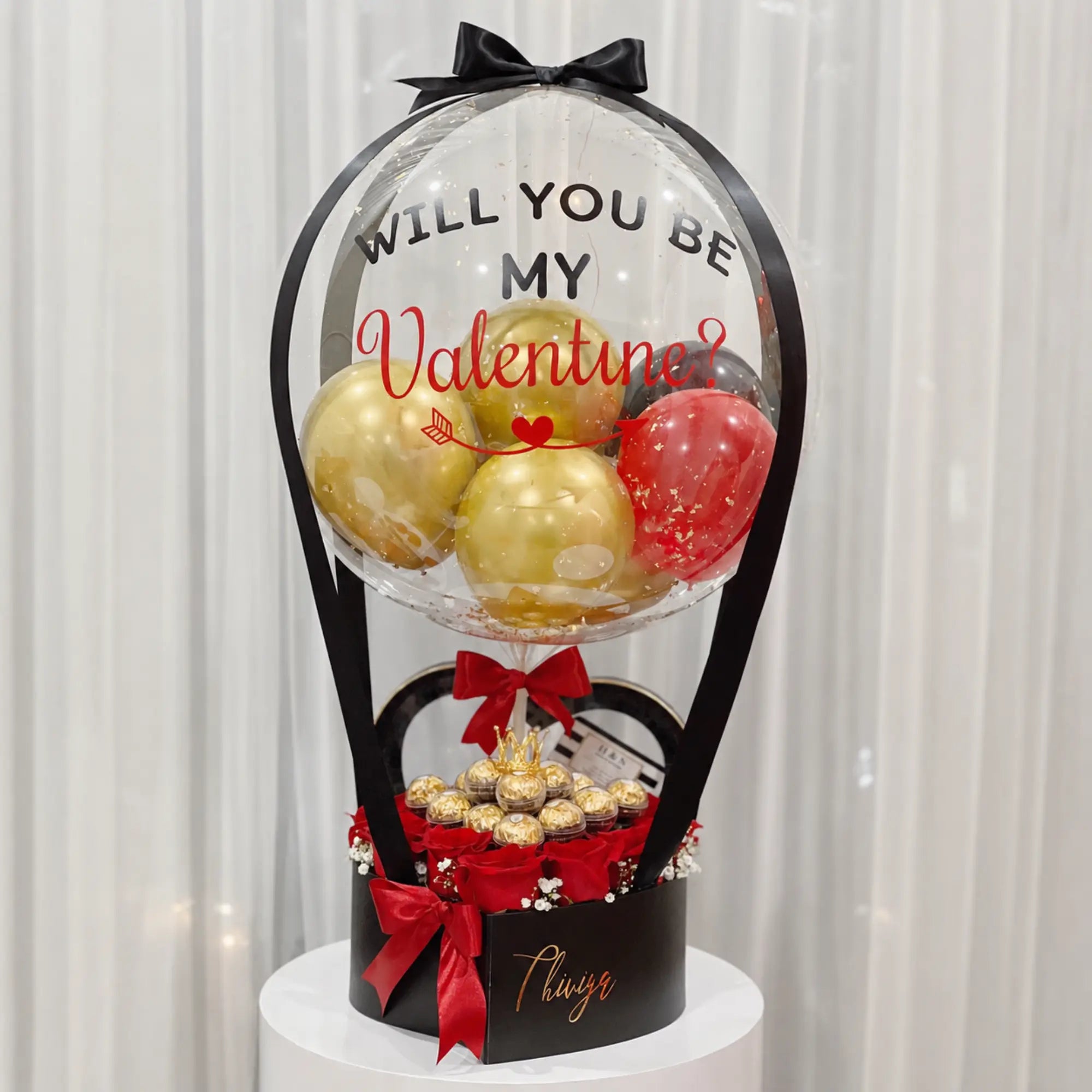 Valentine’s Day proposal gift arrangement featuring a personalized clear “Will You Be My Valentine?” balloon with gold, red, and black balloons, red roses, gold-wrapped chocolates, black gift box, and red ribbon accents.