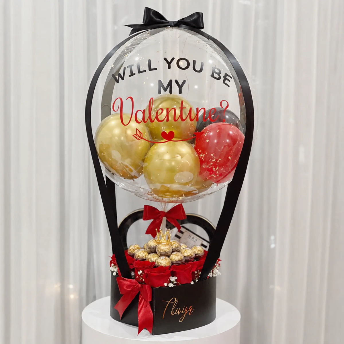 Valentine’s Day proposal gift arrangement featuring a personalized clear “Will You Be My Valentine?” balloon with gold, red, and black balloons, red roses, gold-wrapped chocolates, black gift box, and red ribbon accents.