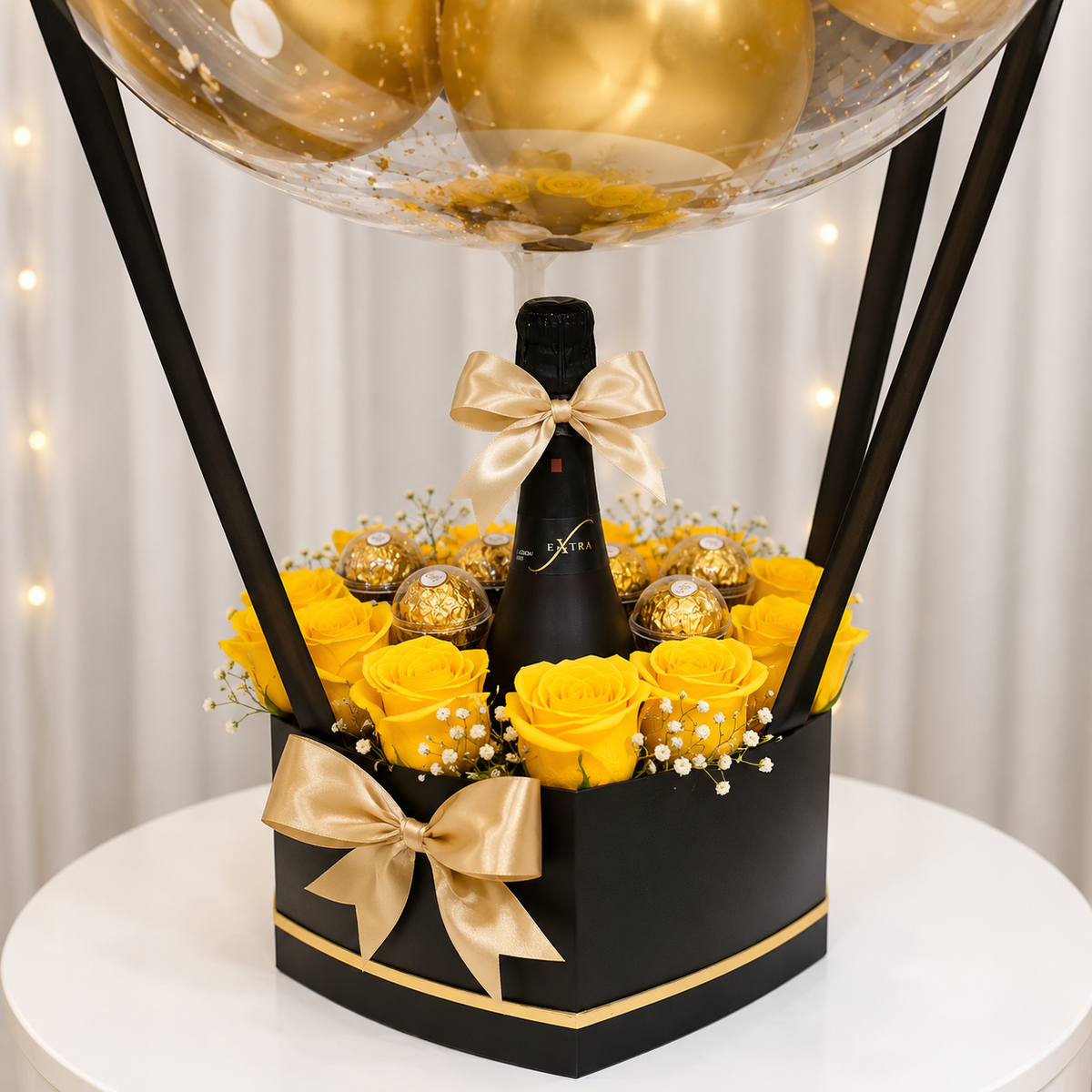 Luxury birthday gift arrangement featuring yellow roses, gold-wrapped chocolates, a black champagne bottle, gold satin bows, and a black and gold gift box beneath a clear balloon.